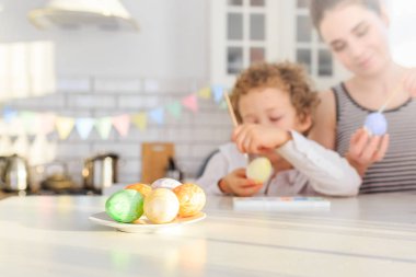 Genç, siyah saçlı mutlu annesi ve kıvırcık sarı oğlu Paskalya için İskandinav tarzında beyaz mutfakta yumurta boyuyorlar.