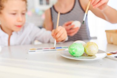 Genç, siyah saçlı mutlu annesi ve kıvırcık sarı oğlu Paskalya için İskandinav tarzında beyaz mutfakta yumurta boyuyorlar.