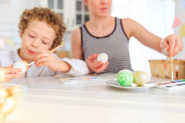 Genç, siyah saçlı mutlu annesi ve kıvırcık sarı oğlu Paskalya için İskandinav tarzında beyaz mutfakta yumurta boyuyorlar.