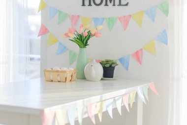 Paskalya evi dekorasyonları, çok renkli bayraklar, laleler ve yumurtalar beyaz bir masada açık bir günde kar beyazı bir mutfakta.