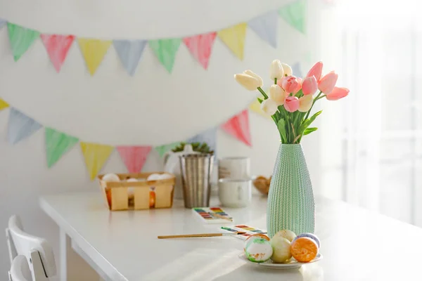 Paskalya evi dekorasyonları, çok renkli bayraklar, laleler ve yumurtalar beyaz bir masada açık bir günde kar beyazı bir mutfakta.