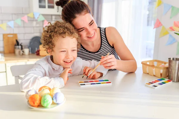 Genç, siyah saçlı mutlu annesi ve kıvırcık sarı oğlu Paskalya için İskandinav tarzında beyaz mutfakta yumurta boyuyorlar.
