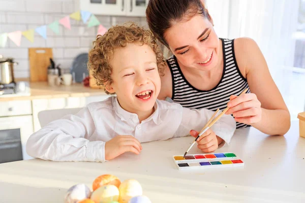 Genç, siyah saçlı mutlu annesi ve kıvırcık sarı oğlu Paskalya için İskandinav tarzında beyaz mutfakta yumurta boyuyorlar.