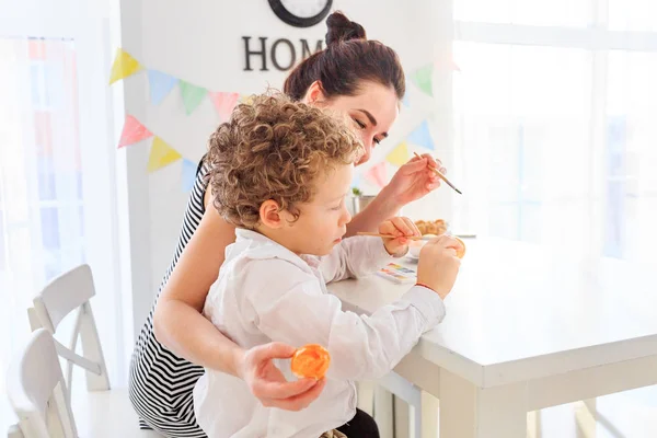 Genç, siyah saçlı mutlu annesi ve kıvırcık sarı oğlu Paskalya için İskandinav tarzında beyaz mutfakta yumurta boyuyorlar.