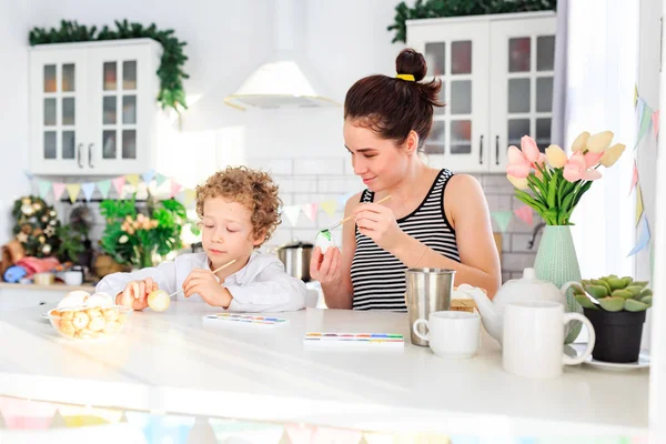 Genç, siyah saçlı mutlu annesi ve kıvırcık sarı oğlu Paskalya için İskandinav tarzında beyaz mutfakta yumurta boyuyorlar.