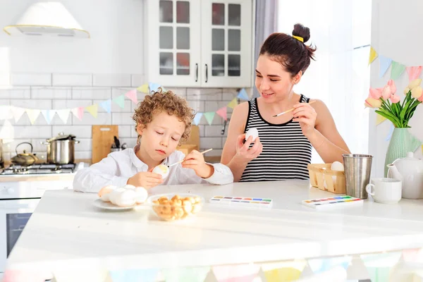Genç, siyah saçlı mutlu annesi ve kıvırcık sarı oğlu Paskalya için İskandinav tarzında beyaz mutfakta yumurta boyuyorlar.