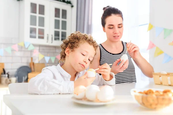 Genç, siyah saçlı mutlu annesi ve kıvırcık sarı oğlu Paskalya için İskandinav tarzında beyaz mutfakta yumurta boyuyorlar.