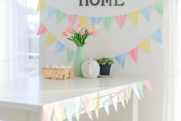 Paskalya evi dekorasyonları, çok renkli bayraklar, laleler ve yumurtalar beyaz bir masada açık bir günde kar beyazı bir mutfakta.