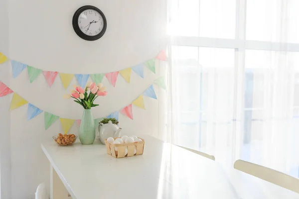 Paskalya evi dekorasyonları, çok renkli bayraklar, laleler ve yumurtalar beyaz bir masada açık bir günde kar beyazı bir mutfakta.