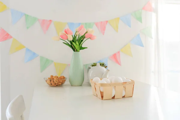 Paskalya evi dekorasyonları, çok renkli bayraklar, laleler ve yumurtalar beyaz bir masada açık bir günde kar beyazı bir mutfakta.