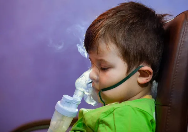 Maskeli küçük bir çocuk, evde nebulizörle solunum yolu tedavisi görüyor. Bebek ağzında nebulizör, astım spreyi ve bronşit tedavisi ile oturuyor.
