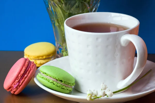 French multicolored cakes macarons and a cup of tea. A bouquet of lily of the valley in a crystal vase. — Stock Photo, Image