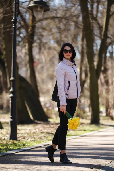 Ein schönes Mädchen mit Sonnenbrille hält einen Strauß Narzissen in der Hand, die im Park spazieren gehen — Stockfoto