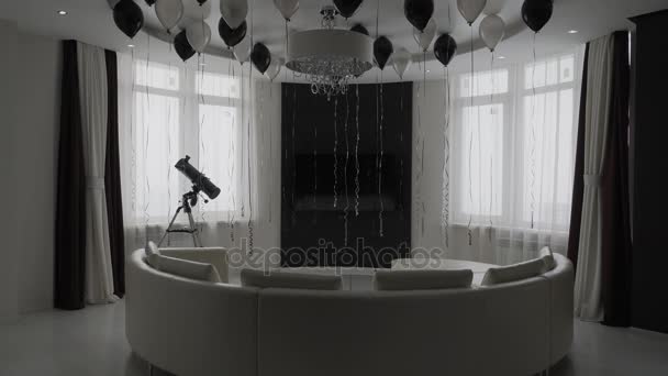 Круглая современная комната в квартире с двумя большими окнами, полупрозрачным кожаным диваном, телевизором и воздушными шарами черного и серебристого цвета — стоковое видео