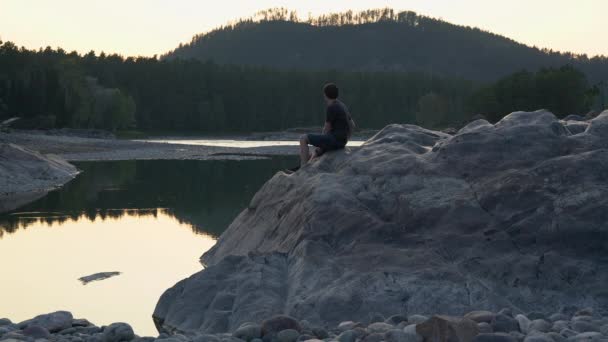 Um homem senta-se em uma grande rocha junto ao lago e olha para a distância para as montanhas ao pôr do sol . — Vídeo de Stock