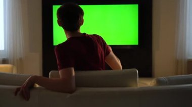 Genç bir adam kanepenin üzerinden atlar ve televizyonu açar ve onu izler. Loş ışık odasında yeşil ekran televizyon.