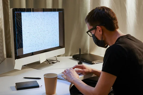 Siyah tıbbi maskeli bir bilgisayar korsanı ya da programcı program kodlu bir monitörün önünde gözlük takıyor..