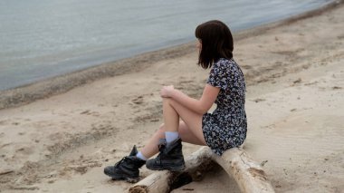 Elbiseli ve çizmeli yalnız bir kız sahilde oturuyor..