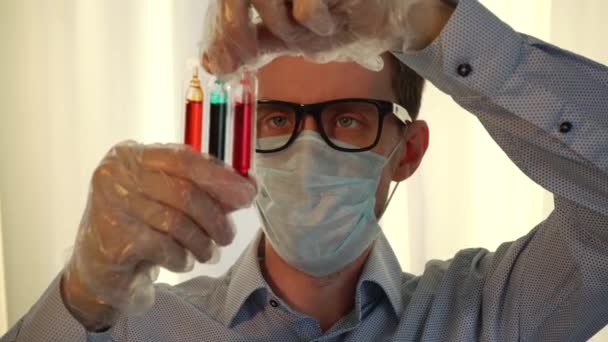 配眼镜和医疗面罩的年轻男性科学家或医生用试管检查彩色样品 — 图库视频影像