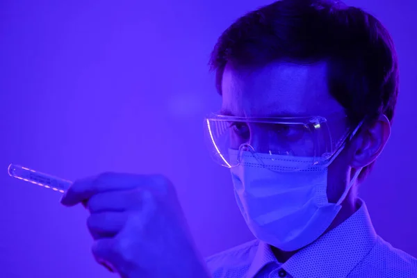 The doctor in glasses and a medical mask looks at the temperature on a thermometer. Portrait in a blue-violet neon light.