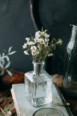 Bahar çiçekleri ve yeşillik dekor cam şişe su ve camına damlacıkları ile tablo içinde. Sıcak gama ve küçük çiçekler ve çim, koyu ahşap arka plan ile ölçülü kompozisyon