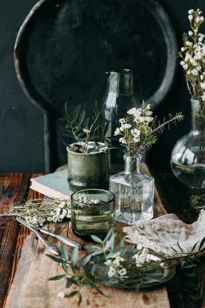 Spring flowers and greens in the decor on the table, in glass bottles with water and droplets on the glass. Warm gamma and restrained composition, with small flowers and grass, on a dark wooden background