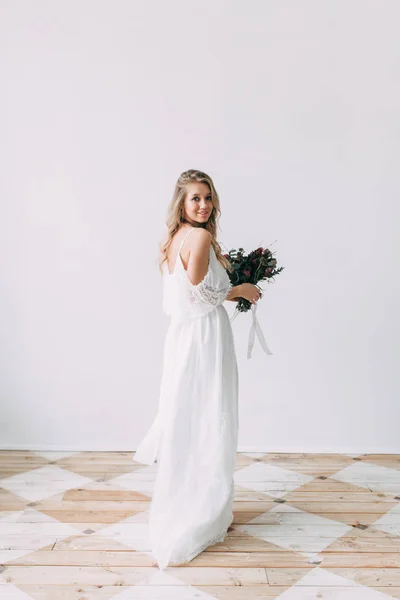 The bride in a light wedding dress with a bouquet of red flowers, on a light wall in the Scandinavian style. European wedding, modern trends and inspiration