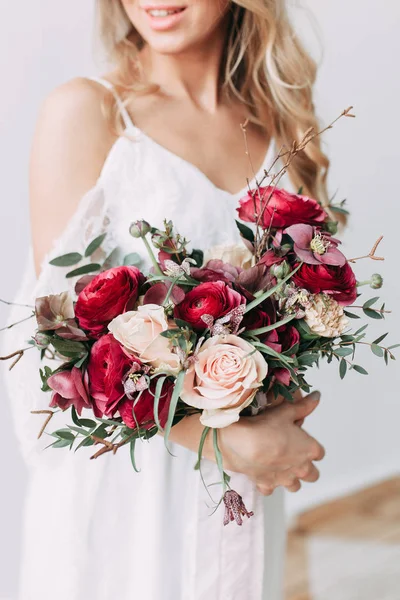 The bride in a light wedding dress with a bouquet of red flowers, on a light wall in the Scandinavian style. European wedding, modern trends and inspiration