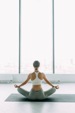  Exercises for self-control in yoga. White Windows with a panorama of the city. Relaxed posture during meditation. The girl is sitting on the Mat, relax.