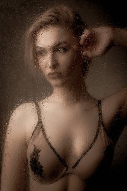 Islak camın arkasında siyah arka planda iç çamaşırlı sarışın bir kız..