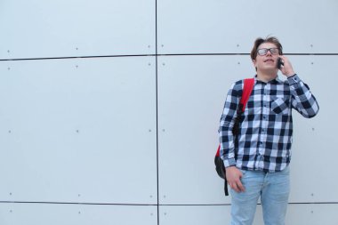 Gözlük, kırmızı sırt çantası ile gülümseyen, telefon, beyaz duvar arka plan üzerinde konuşurken çocuk genç (öğrenci veya öğrenci) gömlek,.