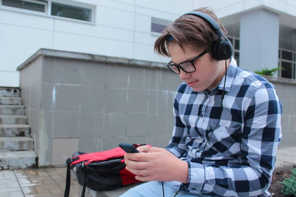 Chlapec dospívající (školák nebo student) v košili, úsměvy v brýlích, poslouchá hudbu na telefon, červený batoh, školní pozadí. — Stock fotografie
