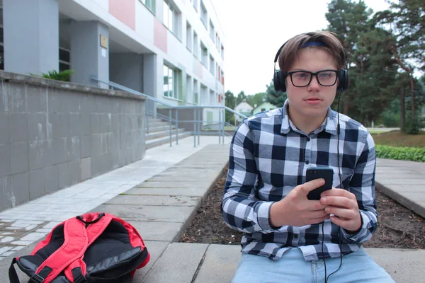 Ragazzo adolescente (scolaro o studente) in camicia, sorride in occhiali, ascolta musica al telefono, zaino rosso, sfondo scolastico . — Foto Stock