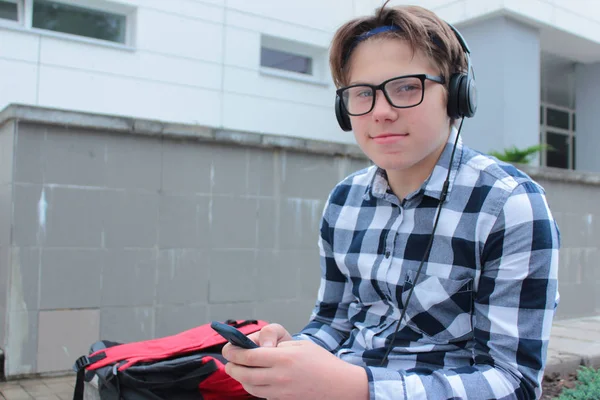 Jongen luistert tiener (scholier of student) in een shirt, glimlacht in glazen, naar muziek op de telefoon, de rode rugzak, de achtergrond van de school. — Stockfoto