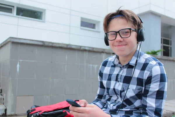 Jongen luistert tiener (scholier of student) in een shirt, glimlacht in glazen, naar muziek op de telefoon, de rode rugzak, de achtergrond van de school. — Stockfoto