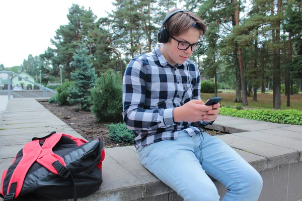 Niño adolescente (escolar o estudiante) en una camisa, sonríe con gafas, escucha música en el teléfono, mochila roja, fondo de la escuela . —  Fotos de Stock