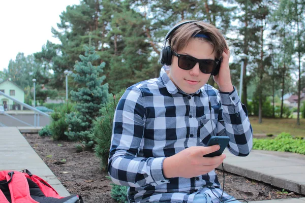 Telefonda, park arka plan müzik dinlemek içinde güneş gözlüğü, gülümseyen çocuk genç (öğrenci veya öğrenci) bir gömlek. — Stok fotoğraf