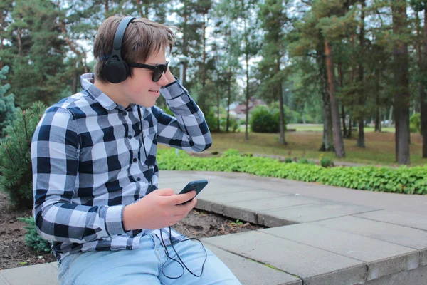 Boy teenager (schoolboy or student) in a shirt, smiling in sunglasses, listening to music on the phone, park background. — Stock Photo, Image