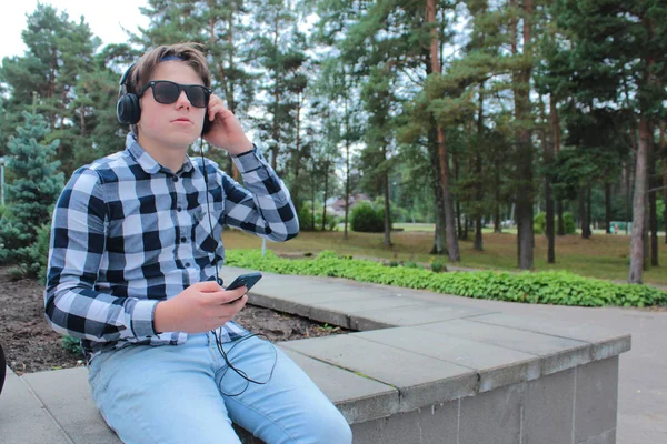 Niño adolescente (colegial o estudiante) en una camisa, sonriendo con gafas de sol, escuchando música en el teléfono, fondo del parque . —  Fotos de Stock