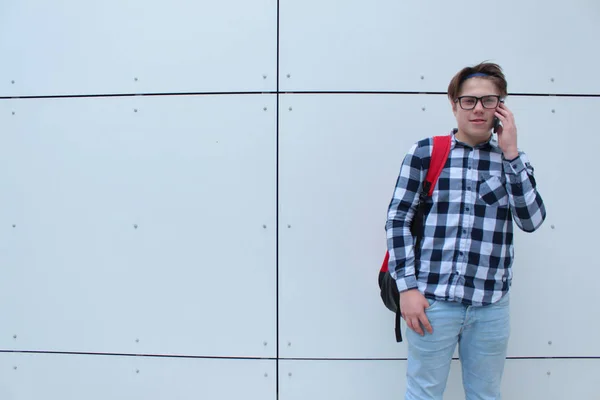 Niño adolescente (colegial o estudiante) en camisa, sonriendo con gafas, mochila roja, hablando por teléfono, fondo blanco de la pared . —  Fotos de Stock