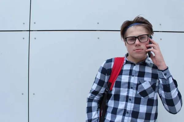 Adolescent garçon (écolier ou étudiant) en chemise, souriant avec des lunettes, sac à dos rouge, parler au téléphone, fond de mur blanc . — Photo