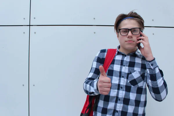 Chlapec dospívající (školák nebo student) v košili, s brýlemi, červený batoh úsměvem, mluví na pozadí telefonu, bílé zdi. — Stock fotografie