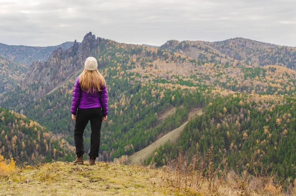 A girl in a lilac jacket standing on a mountain, a view of the mountains and an autumn forest by a cloudy day