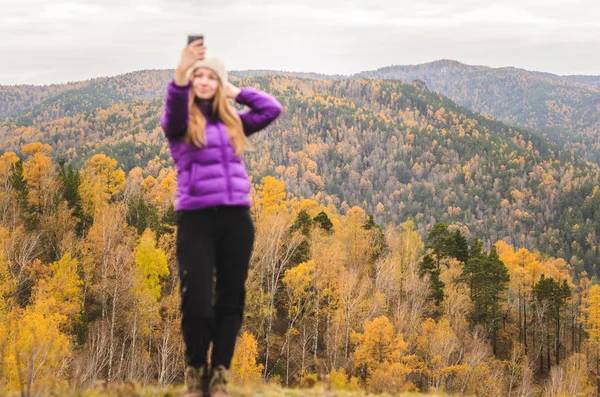 A girl in a lilac jacket makes a salfi on a mountain, a view of the mountains and an autumnal forest by a cloudy day, free space for text