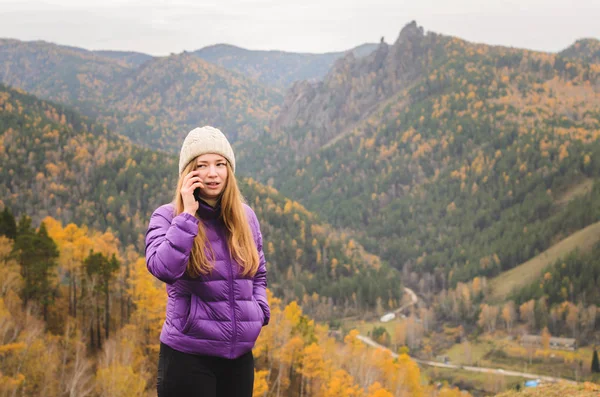 A girl in a lilac jacket talking on the phone in the mountains, an autumn forest with a cloudy day, free space for text