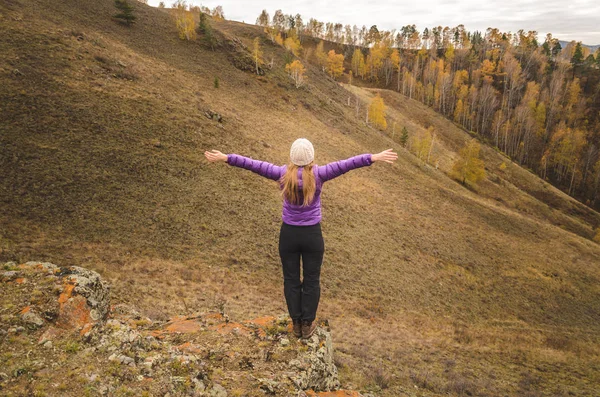 A girl in a lilac jacket stretching her arms on a mountain, a view of the mountains and an autumn forest by a cloudy day, free space for text