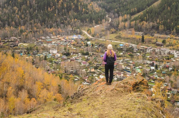 A girl in a lilac jacket looks out into the distance on a mountain, a view of the mountains and an autumnal forest by an overcast day. Free space for text