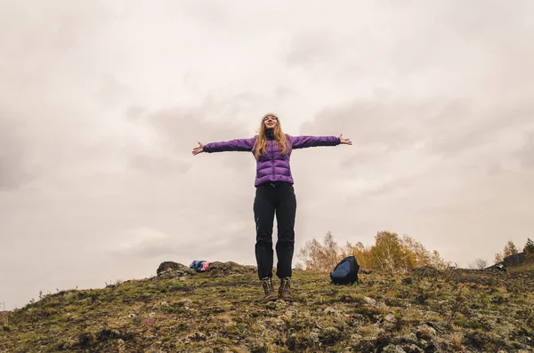 A girl in a lilac jacket stretching her arms on a mountain, a view of the mountains and an autumn forest by a cloudy day, free space for text