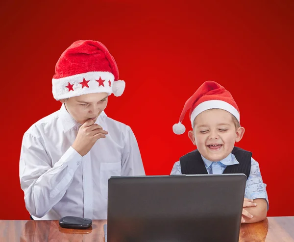 Enfants en casquettes Père Noël regardant un ordinateur portable sur un fond rouge — Photo