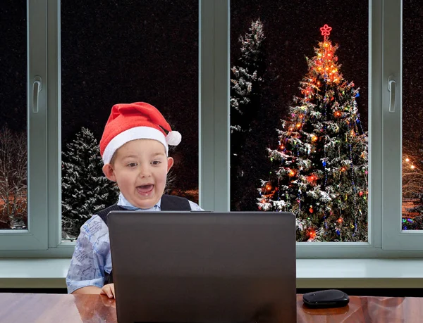 Le garçon regarde un ordinateur portable contre la fenêtre suivi d'un sapin de Noël — Photo